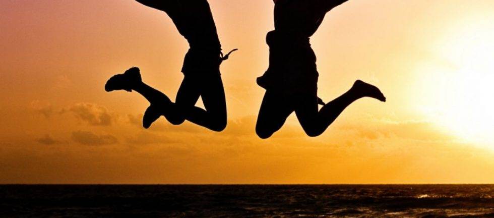 Two silhouetted figures joyfully leap against a vibrant sunset over the ocean.