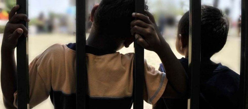 Two children stand behind bars, looking forlorn. Child poverty, vulnerable children.