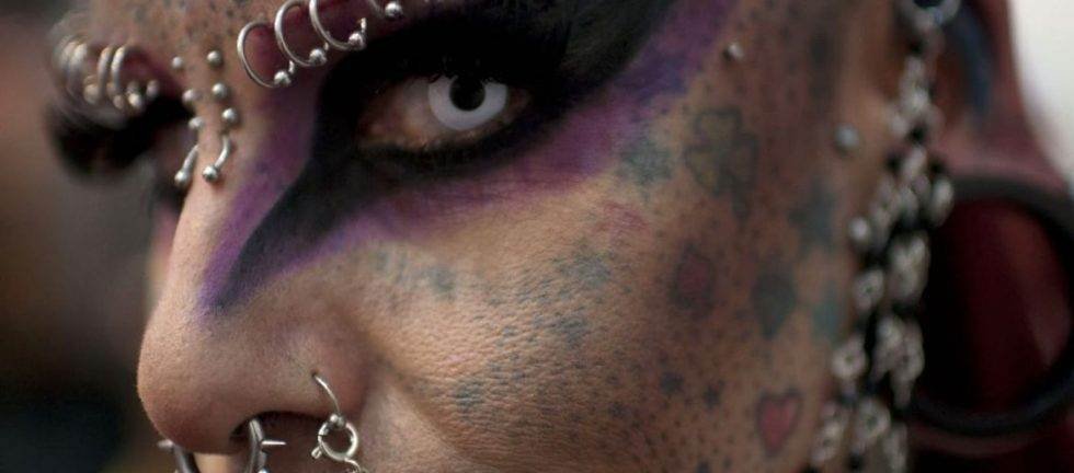 Close-up of a heavily pierced and tattooed face with vibrant purple eye makeup.