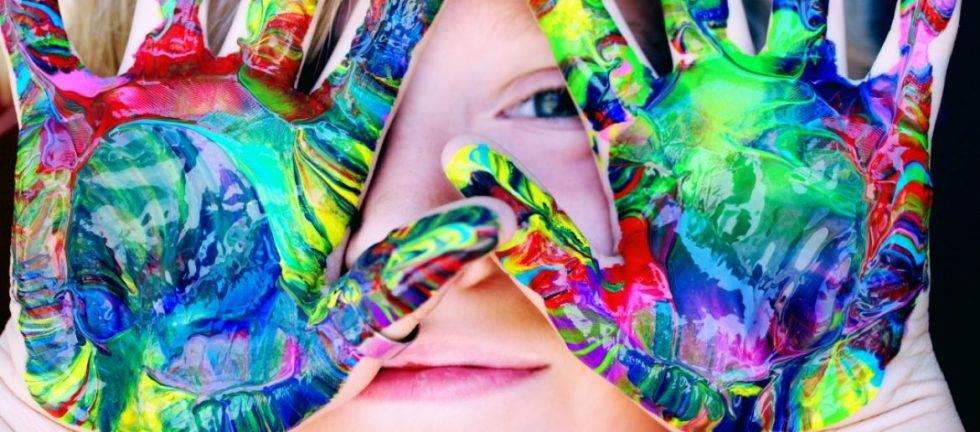 Child's face framed by brightly colored, paint-covered hands