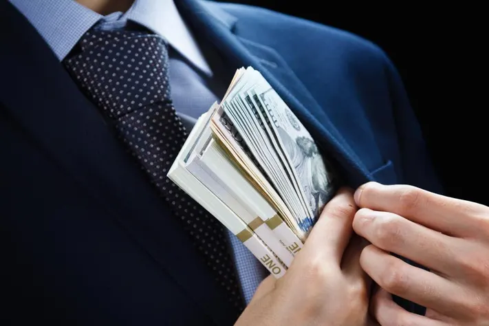 Close-up of a person's hands concealing a large stack of cash in a suit jacket pocket.