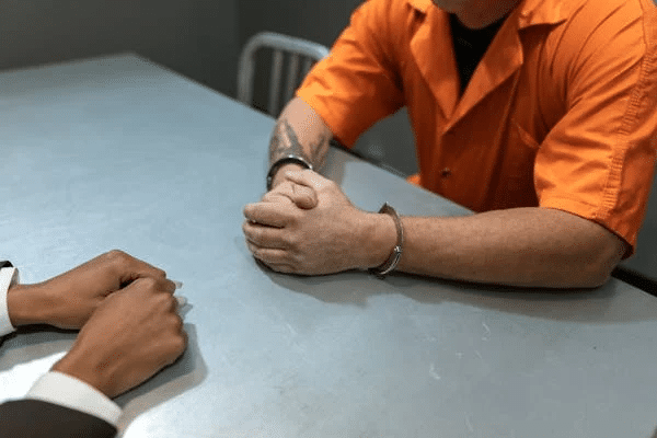 A prisoner in handcuffs sits across a table from a lawyer, discussing plea bargaining options in NSW.