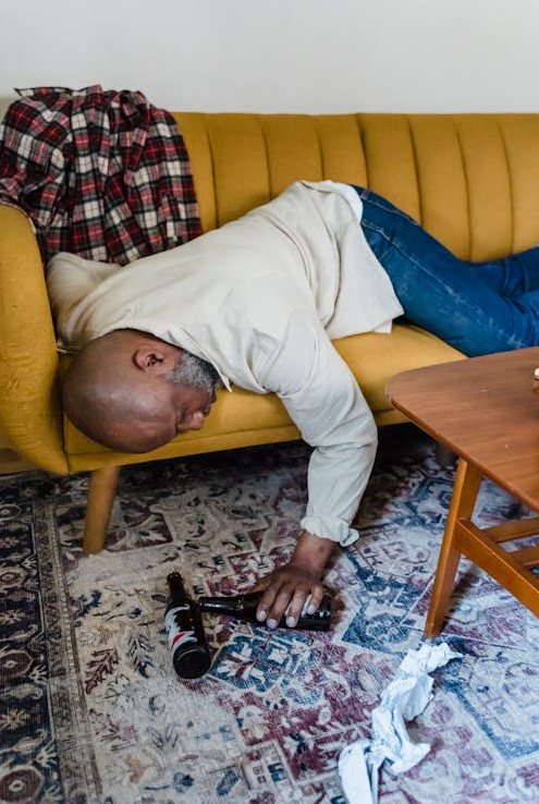 Man slumped over a yellow couch reaching for an empty bottle, illustrating intoxication as a criminal defence concept.