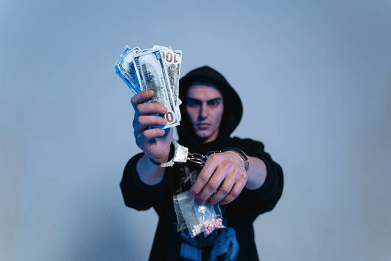 A hooded man in handcuffs holds cash and a bag suggesting blackmail and extortion offences in NSW.