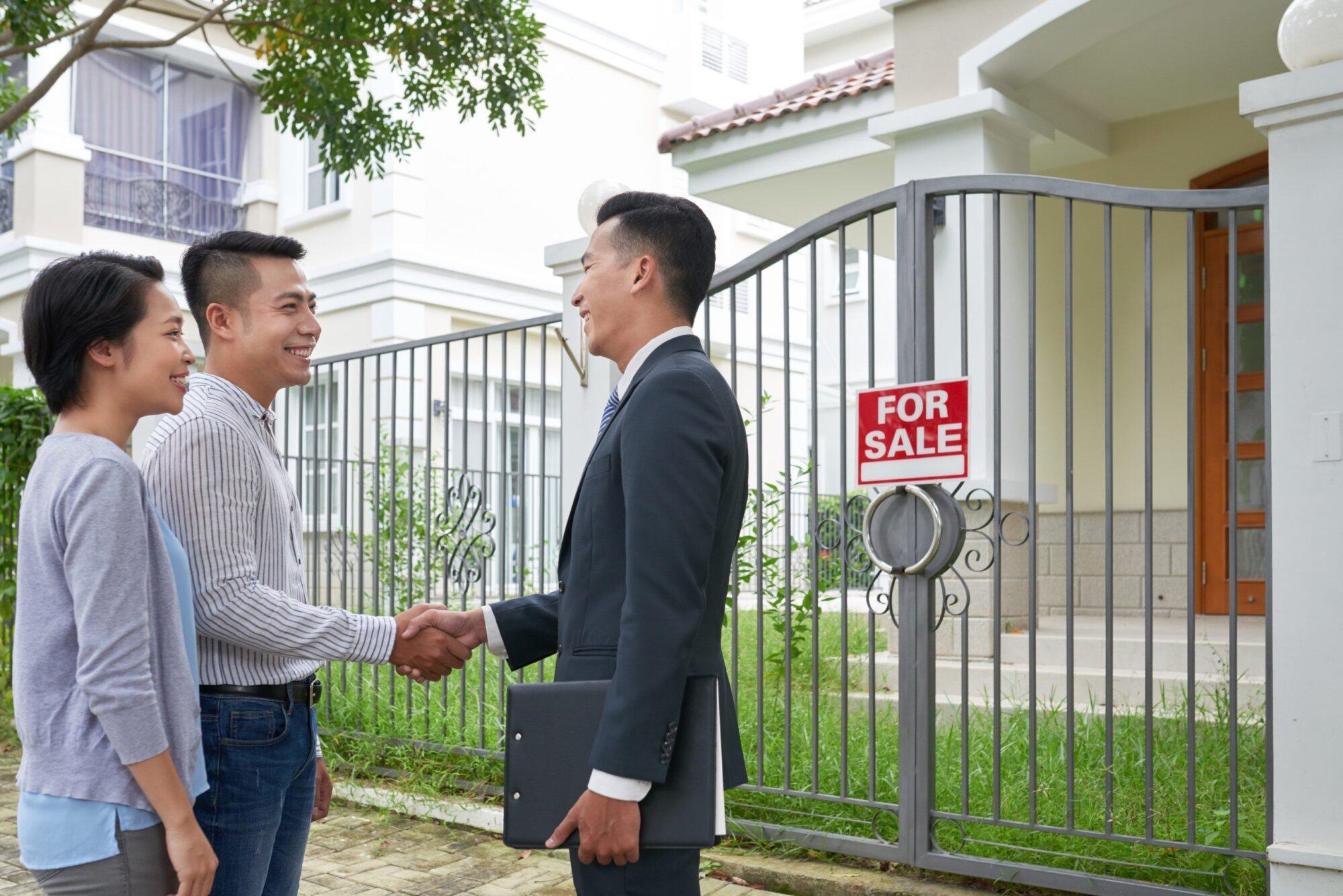 Couple shaking hands with realtor outside a house with a "For Sale" sign, relevant to property offences NSW context.