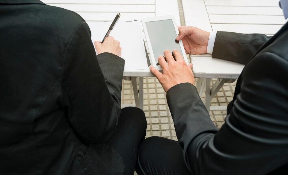 Two people in suits discuss withdrawing from a joint criminal enterprise while sitting at a table with a tablet.