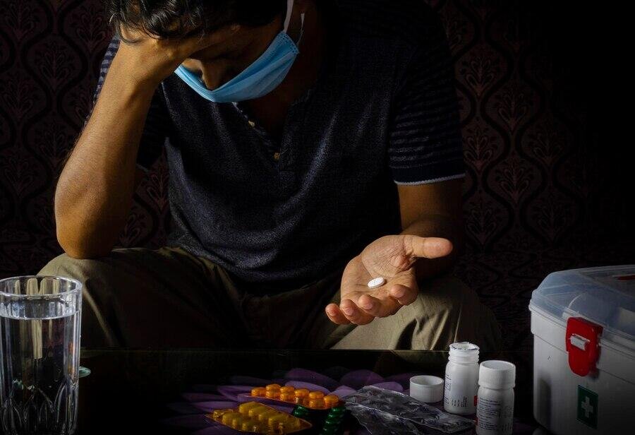 A man wearing a mask holds a pill in one hand, with medicine bottles and a white box nearby, suggesting drug misuse.