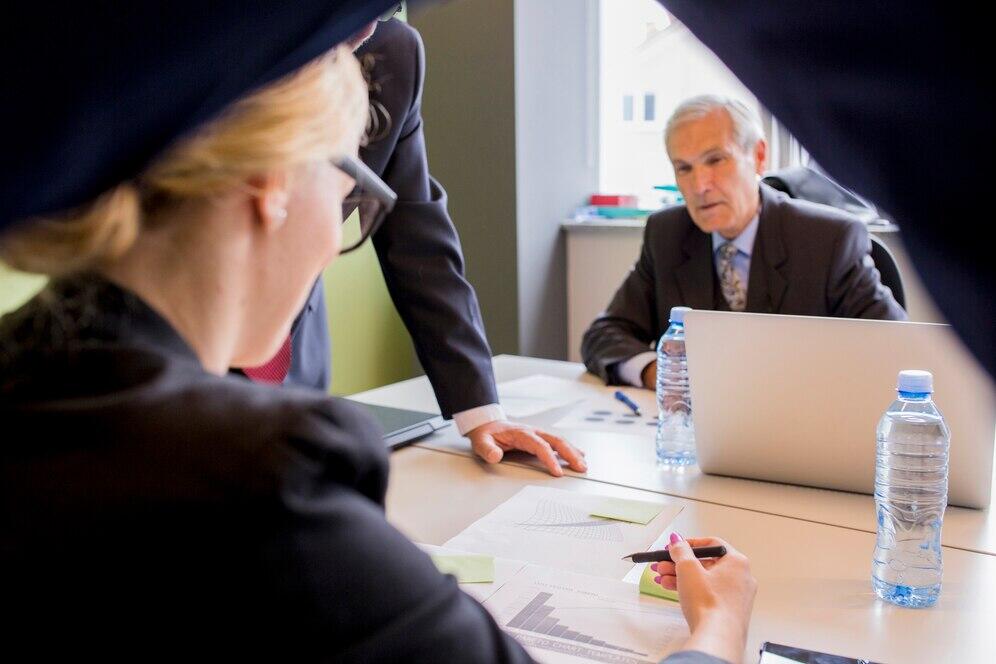 Business meeting discussing joint criminal enterprise, with documents, laptops, and water bottles on the table.