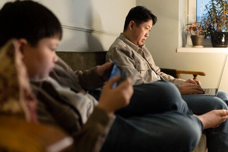 Father and son relax on a couch, one using a laptop, the other a phone.