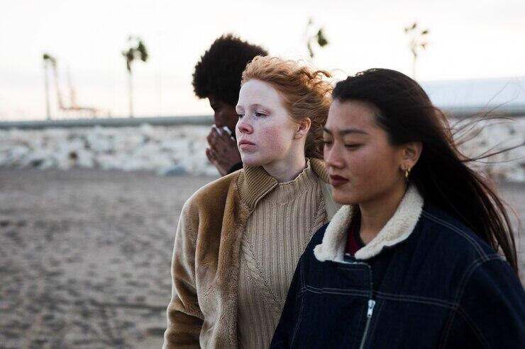 Three young women stand together on a beach. One woman is wearing a tan sweater and brown jacket. Another woman wears a dark denim jacket.