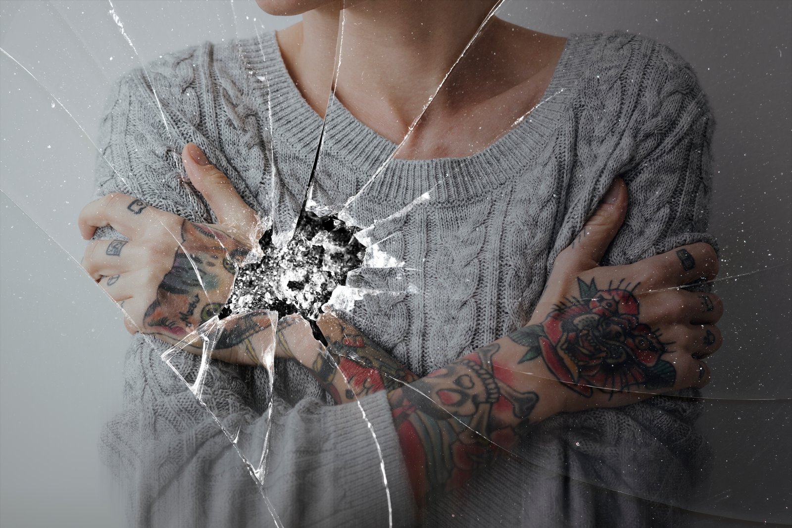 Person with tattoos hugging themselves, superimposed on cracked glass.