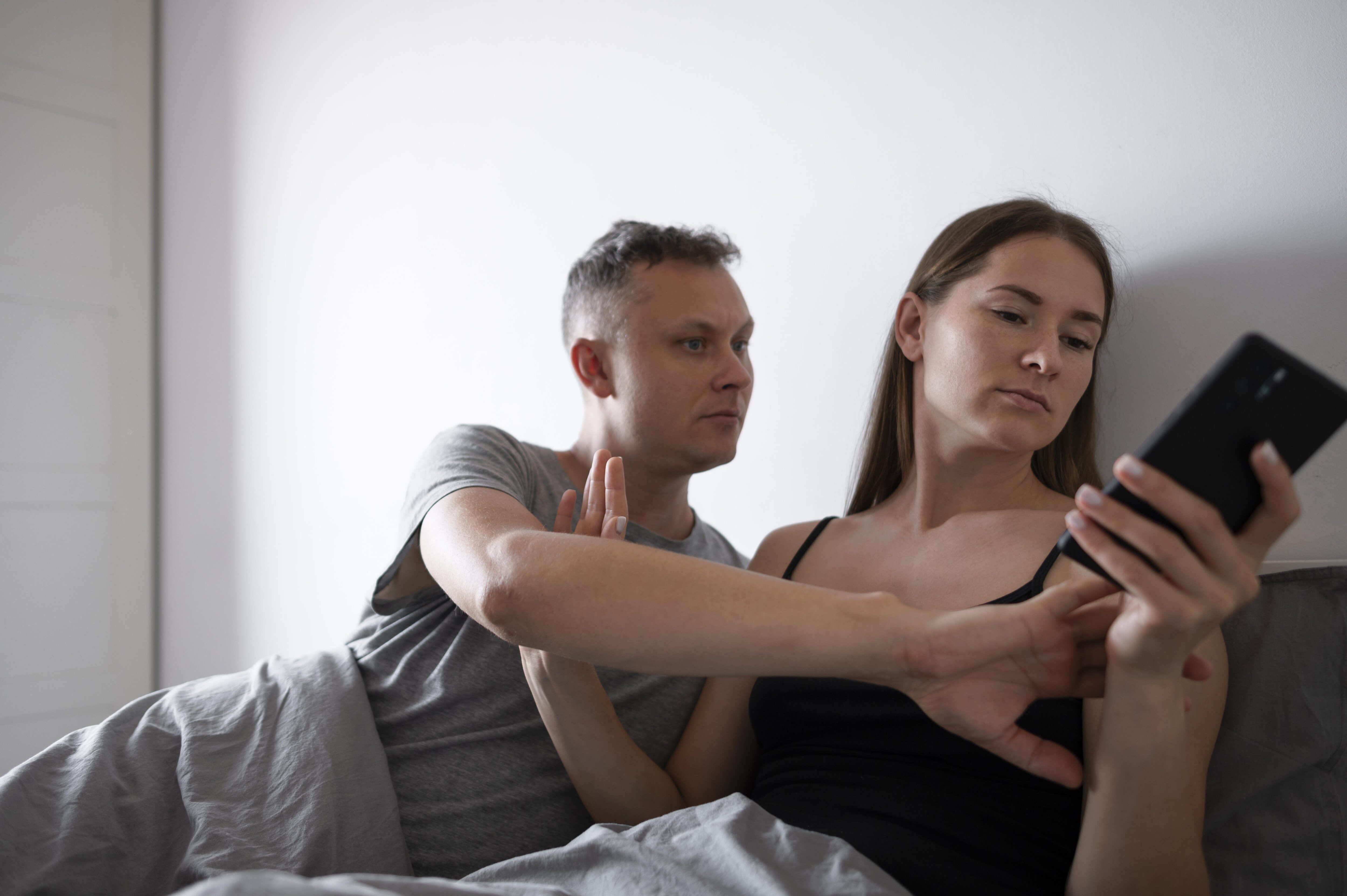 Couple in bed, woman on phone, man looking concerned.